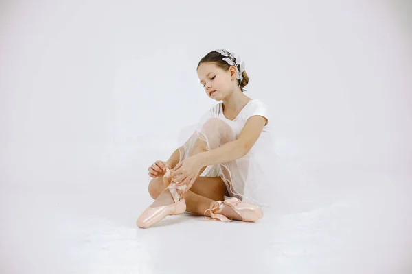 Little ballerina on white background. Brunette girl wearing white ballet suit. Girl sitting on a floor and wearing pointe shoes.