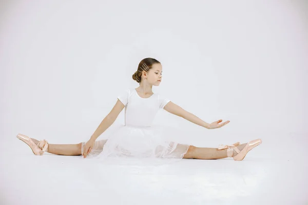 Little ballerina on white background. Brunette girl wearing white ballet suit. Girl sitting on a twine.