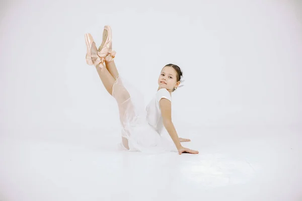 Little ballerina on white background. Brunette girl wearing white ballet suit. Girl sitting on a floor.