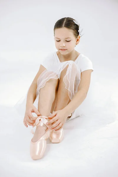 Little ballerina on white background. Brunette girl wearing white ballet suit. Girl sitting on a floor and wearing pointe shoes.