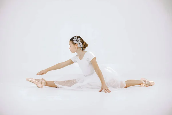 Little ballerina on white background. Brunette girl wearing white ballet suit. Girl sitting on a twine.