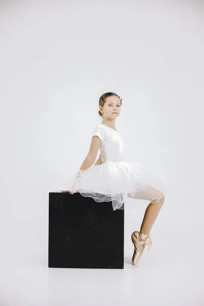 Little ballerina on white background. Brunette girl wearing white ballet suit. Girl sitting on black cube.