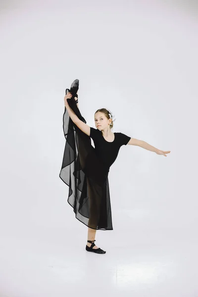 Little ballerina on white background. Brunette girl wearing black ballet suit. Girl doing gymnastic excercises.