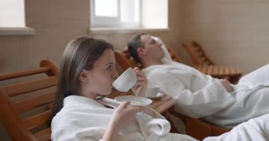 Young lovers in bathrobes are relaxing in chairs in spa salon, drinking tea, talking. Wellness, romantic relationship and happiness concept.