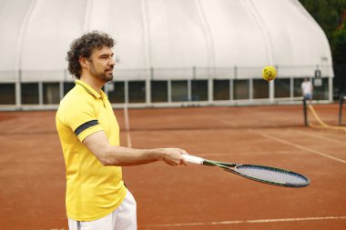 Portrait of tennis player man, front view. Man standing on a court and holding a racquet. Man wearing yellow t-shirt and white shorts.