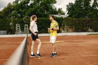 Curly man and woman tennis players standing on open summer court with rackets in hands. Couple friendly chatting before the match. Man wearing yellow t-shirt and woman white one.