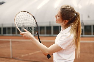 Woman tennis player standing on open summer court with racket in hand. Woman looking at her racket. Woman wearing white t-shirt.