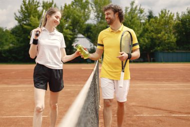 Curly man and woman tennis players standing on open summer court with rackets in hands. Couple friendly chatting before the match. Man wearing yellow t-shirt and woman white one.