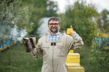 Experienced senior apiarist in his apiary setting a fire in a bee smoker. Beekeeper harvesting honey. Bee smoker is used to calm bees before frame removal.