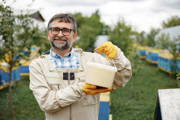 Beekeeper holding bucket of honey in hands. Farmer wearing bee suit working with honeycomb in apiary. Man looking at camera.