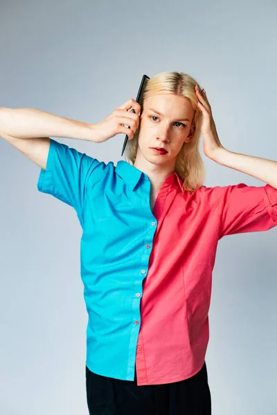 Young blond man has make up on one half of his face. Man wearing fashionable shirt divided on two colours bright pink and blue. Man isolated on grey background.