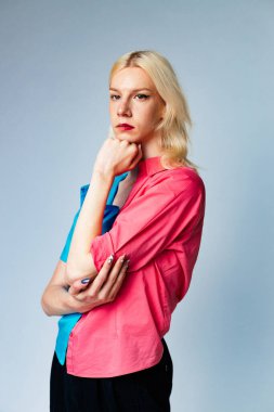 Young blond man has make up on one half of his face. Man wearing fashionable shirt divided on two colours bright pink and blue. Man isolated on grey background.