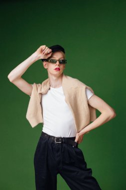 Young blond man with make up on his face and sunglasses. Man wearing fashionable white t-shirt and black cap. Man isolated on green background.