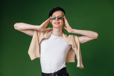 Young blond man with make up on his face and sunglasses. Man wearing fashionable white t-shirt and black cap. Man isolated on green background.