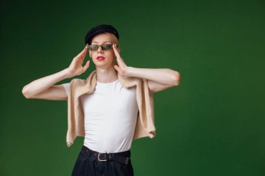 Young blond man with make up on his face and sunglasses. Man wearing fashionable white t-shirt and black cap. Man isolated on green background.