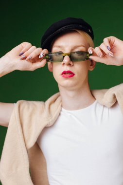 Young blond man with make up on his face and sunglasses. Man wearing fashionable white t-shirt and black cap. Man isolated on green background.