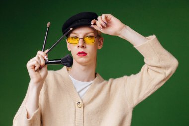 Young blond man with make up on his face wearing sunglasses. Man wearing fashionable beige sweater and black cap. Man isolated on green background and holding brushes for make up in hands.