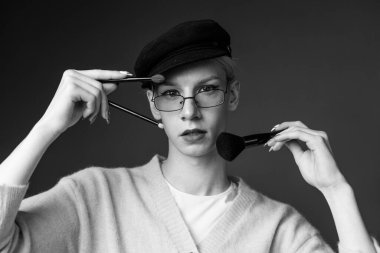 Young man with make up on his face wearing sunglasses. Man wearing fashionable sweater and cap. Man holding brushes for make up in hands.