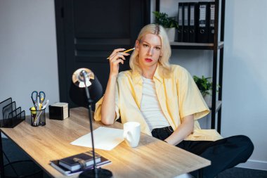 Man with make up on his face sitting in office. Man wearing fashionable white t-shirt and yellow shirt. Man has long blonde hair.