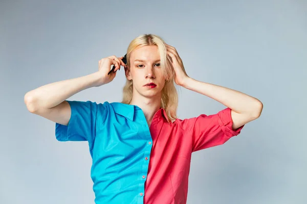 Young blond man has make up on one half of his face. Man wearing fashionable shirt divided on two colours bright pink and blue. Man isolated on grey background.