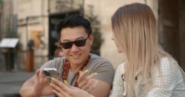 Multiracial couple travelling. Sitting in the city square and using smartphone, looking for tourists spots.