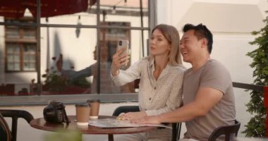 Happy young tourist couple taking selfies and enjoying holidays at street cafeteria. Asian man and Caucasian woman.