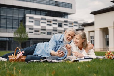 Yazın çimlerin üzerinde battaniyeye uzanmış beyaz yaşlı bir çift. Kadın ve erkek telefona bakıyor. Beyaz tişörtlü kadın ve mavi erkek..