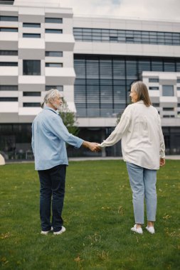 Caucasian elderly couple standing on a grass in summer. Man and woman holding hands. Woman wearing white shirt and man blue one.