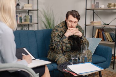 Terapi sırasında koltukta oturan asker ve psikiyatristin kesilmiş fotoğrafı. Askeri üniforma giyen adam. Psikiyatristle konuşan erkek savaşçı..