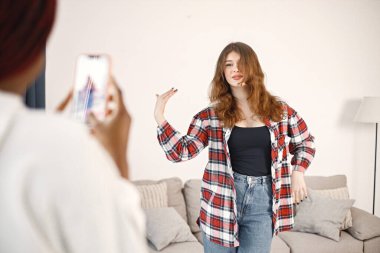 Poz veren beyaz bir kıza odaklan. Evde ekonomi sınıfının yanında duran iki genç kadın. Siyah kız beyaz kapüşonlu, beyaz kız ekose gömlek giyiyor..