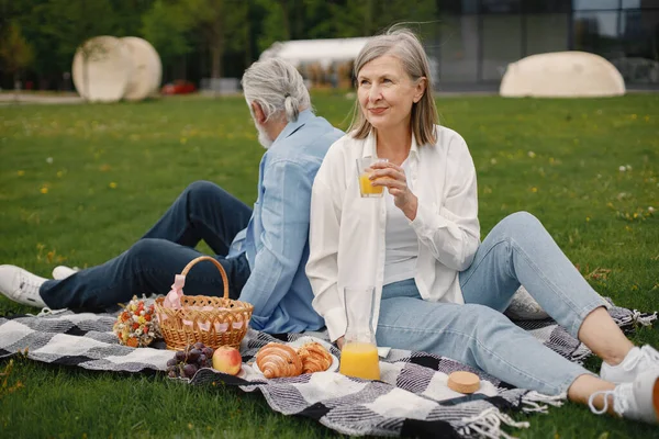 Beautiful caucasian elderly couple sitting on a grass in the park in summer. Man and woman sitting on blanket at the park drinking juice and sharing few precious memories. Woman wearing white shirt