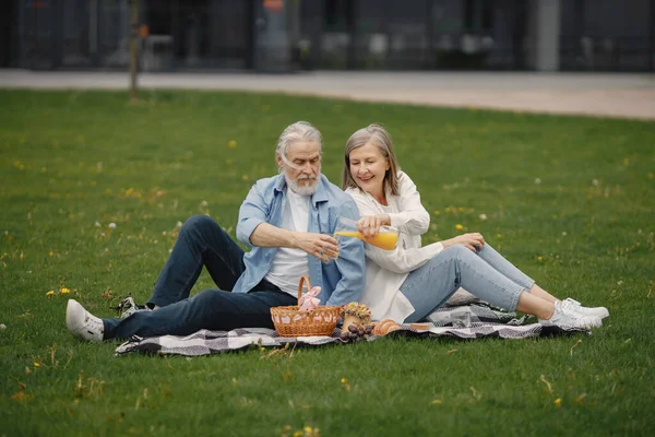 Beautiful caucasian elderly couple sitting on a grass in the park in summer. Man and woman sitting on blanket at the park drinking juice and sharing few precious memories. Woman wearing white shirt