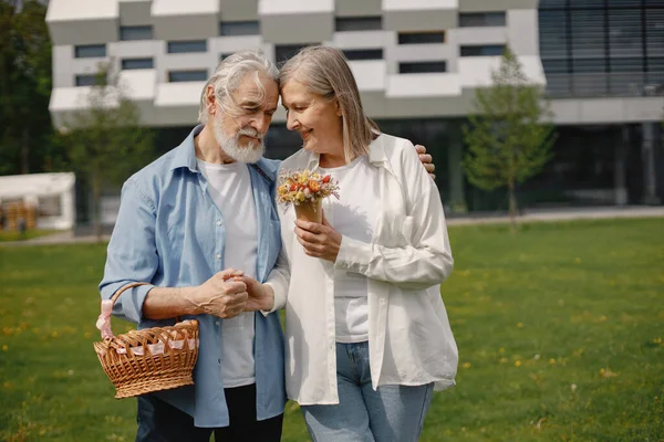 Caucasian elderly couple standing on a grass in summer. Man holding straw basket and woman flowers. Woman wearing white shirt and man blue one.