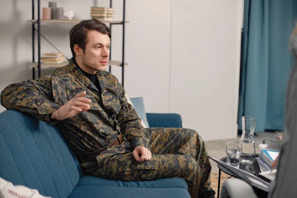 Cropped photo of a soldier sitting on couch during therapy session. Man wearing military uniform. Male warrior with ptds talking to psychiatrist.