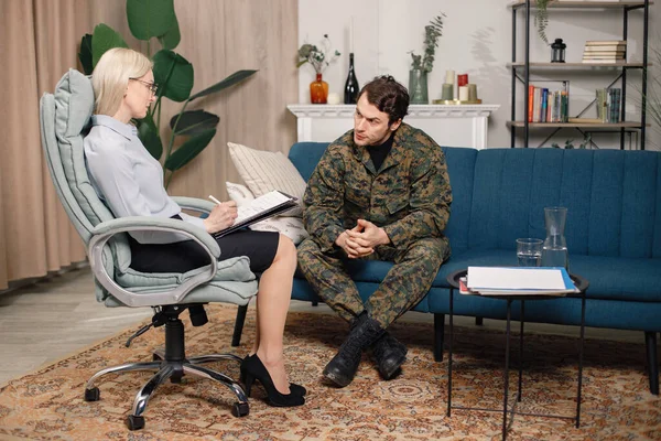Soldier and psychiatrist sitting on couch during therapy session. Man wearing military uniform. Male warrior with ptds talking to psychiatrist.
