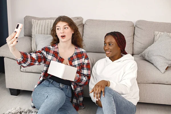 Two young woman sitting on a floor near sofa and make a selfie on a smartphone. Black girl wearing white hoodie, caucasian girl wearing plaid shirt. Girls holding a carton box gift.