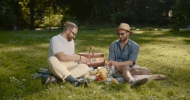Man spending weekend time with boyfriend at summer picnic. Handsome bearded gay couple sitting on blanket and talking at date. Romance concept, lgbtq concept.