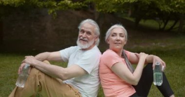 Smiling senior couple in park exercising taking a break hydrating. Healthy lifestyle , retirement.