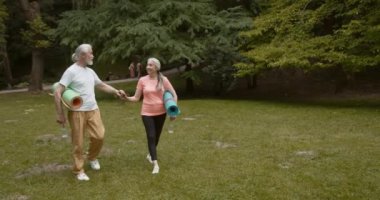 Spor antrenmanından sonra spor paspasları taşıyan mutlu yaşlı çift. Yaşlı karı koca el ele yoga minderleri tutuyor, park yolunda birlikte yürüyor, birbirlerine bakıyor ve konuşuyorlar.