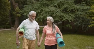 Spor antrenmanından sonra spor paspasları taşıyan mutlu yaşlı çift. Yaşlı karı koca el ele yoga minderleri tutuyor, park yolunda birlikte yürüyor, birbirlerine bakıyor ve konuşuyorlar.