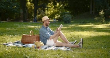 Summertime, weekend concept. Man in a straw hat sitting on a blanket, smiling and reading a book. Man on a picnic outdoors.