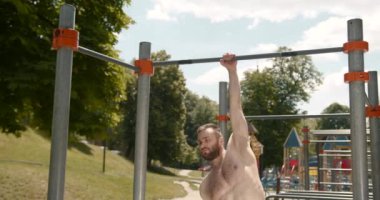 Young bearded man is doing pull ups exercises on horizontal bar outdoors. Guy is training alone in city park on summer day. Active person.