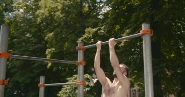 Young bearded man is doing pull ups exercises on horizontal bar outdoors. Guy is training alone in city park on summer day. Active person.