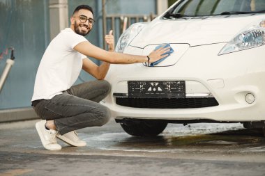 Young man in fashion clothes wiping his car with a rug after washing at car wash station. Man wearing white t-shirt and jeans. Indian man looking at camera and show sign like.