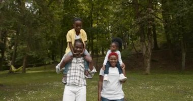Black family walking on the weekend in te green park. Cheerful parents carrying kids on shoulders.