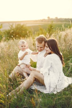 Photo of a young family sitting at the field on a sunny day. Brunette mother, father and their little blonde son posing for a photo. Man and woman looking at their son.