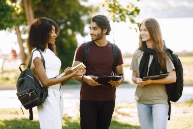 Group of smiling international students standing together in park at university. African and caucasian girls and indian boy talking outdoors. Three friends disscussing about education.