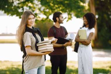 Focus on a smiling caucasian girl who standing sepately. Group of international students standing together in park at university. Blurred african idian boy talking outdoors.