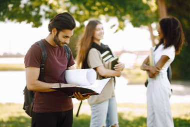 Focus on a indian boy who standing sepately. Group of international students standing together in park at university. Blurred african and caucasian girls talking outdoors.
