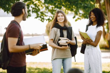 Group of international students standing together in park at university. African and caucasian girls and indian boy talking outdoors. Three friends disscussing about education.
